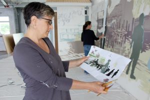 Architect and artist Mary-Ann Agrestic shows her drawing of the Sea Captain's Row mural to be installed on Pleasant Street in Hyannis, as artist Jackie Reeves paints in the background.